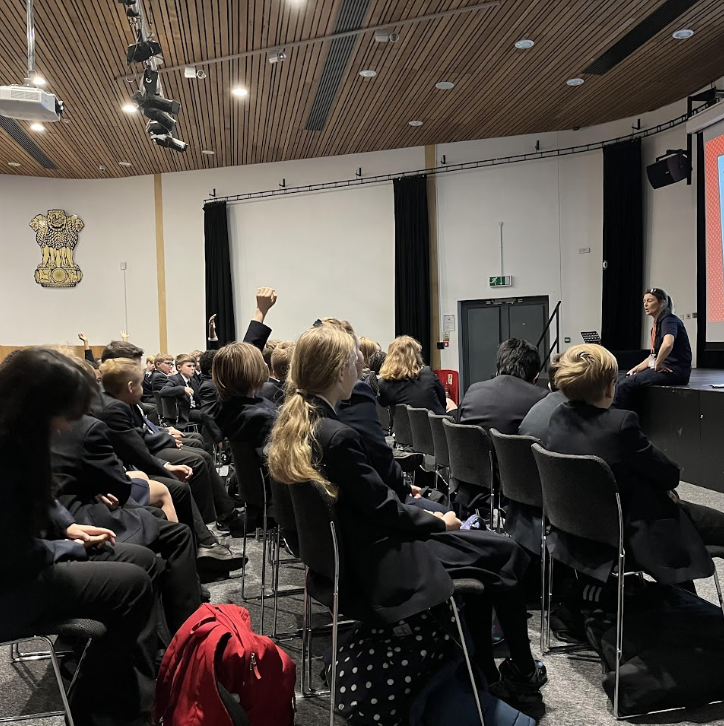 A large group of Leigh Academy students can be seen sat on chairs in the main hall area, listening to a speaker on the stage at the front of the room.