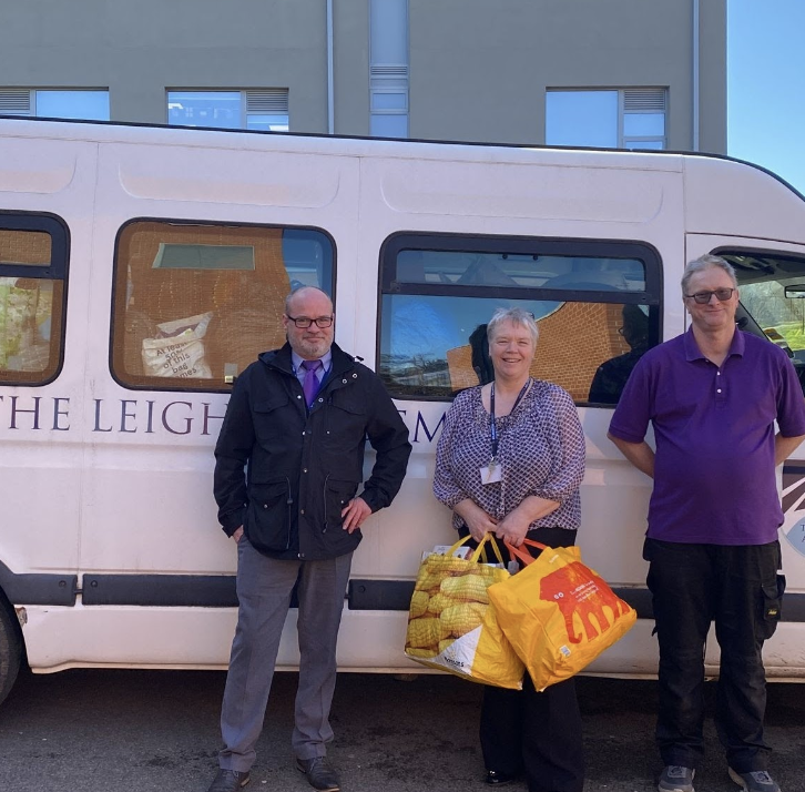 Three staff members are pictured standing beside the academy's minibus smiling. The lady in the centre is holding two bags full of items.