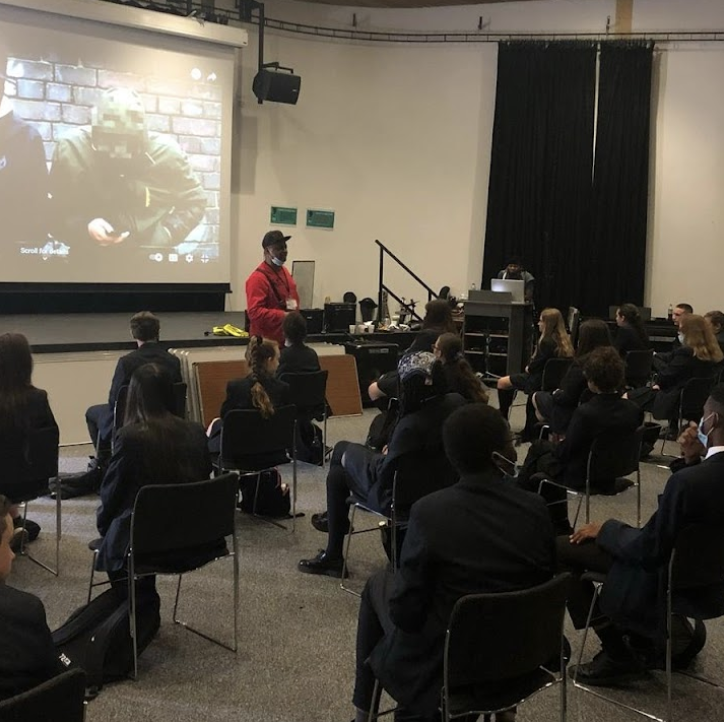 Students are pictured sitting down on chairs in the main hall, listening to a speaker at the front of the room.