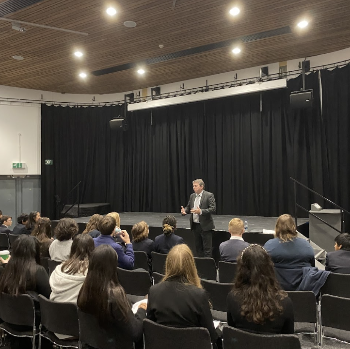 A large group of Leigh Academy students are pictured seated in the main hall, listening to a speaker at the front of the room.