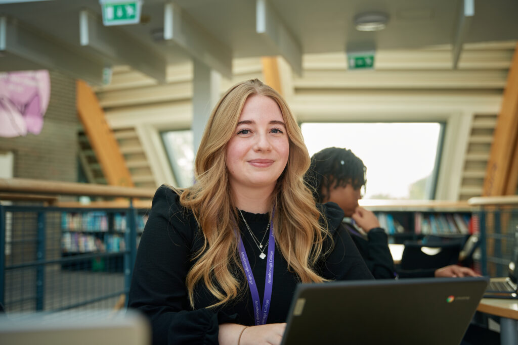 A female Post-16 student is seen smiling for the camera whilst working on a Chromebook laptop in an open area of the academy building.