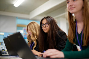 Three girls from Post-16 are shown sat at their desks with laptops in front of them, looking towards the front of the classroom.