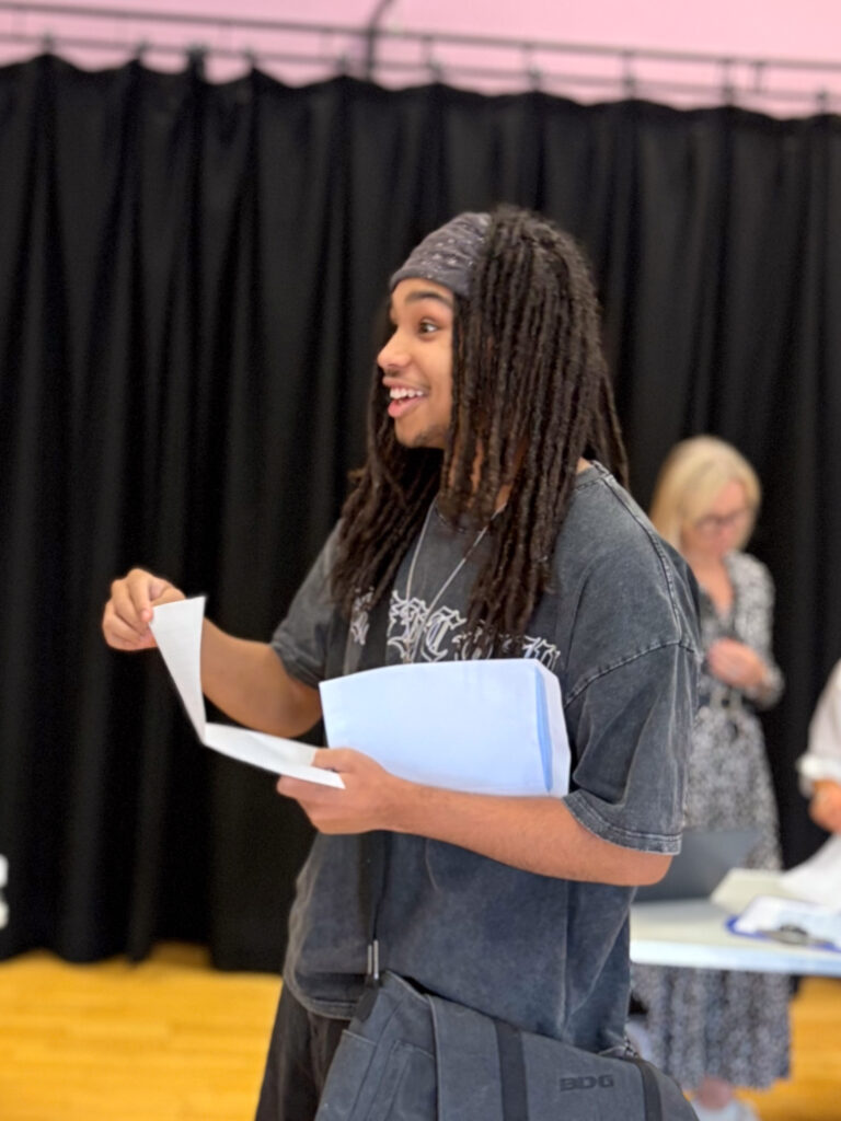 A young man with dreadlocks and a bandana is holding papers (likely results) and a white envelope. He's looking to the side with an open-mouthed, excited expression, as if speaking to someone or reacting