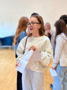 A young woman wearing glasses and a light-colored sweatshirt is holding papers. she's smiling broadly and looking to the side, seemingly happy with what she's seeing.