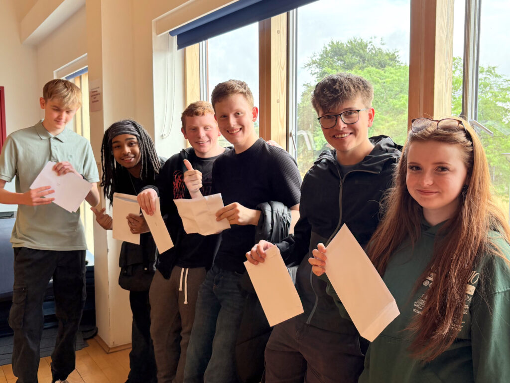 A group of six young people are standing in a row, most holding papers. They are all smiling and looking towards the camera, suggesting they are happy with their results. One individual in the center is giving a thumbs-up.