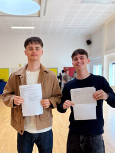 Two young men are standing next to each other, both holding up sheets of paper that also look like official result documents from the "International Baccalaureate Organisation." They are both smiling and looking at the camera.