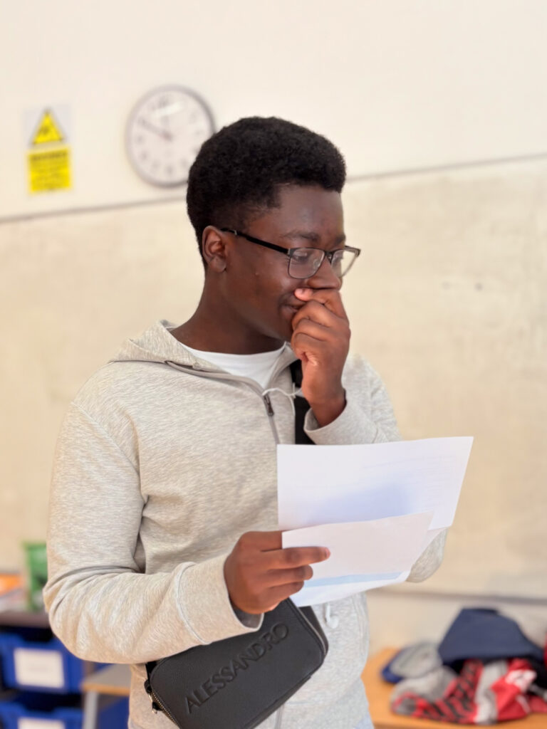 A young man wearing glasses and a grey hoodie is holding papers and looking down at them, with one hand covering part of his mouth, indicating a thoughtful or perhaps surprised reaction.