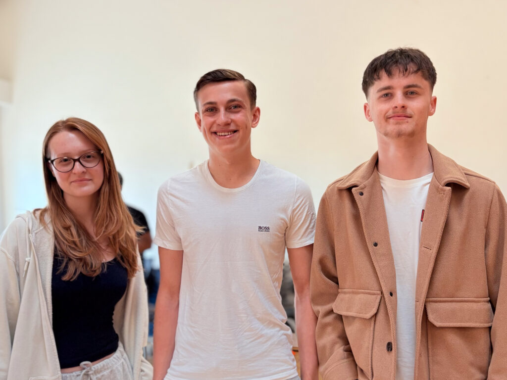 Three young people are standing side-by-side, looking at the camera. The young woman on the left has glasses and long hair, the young man in the middle is smiling broadly and wearing a white t-shirt, and the young man on the right has short dark hair and is wearing a brown jacket. They are posing for a group photo.
